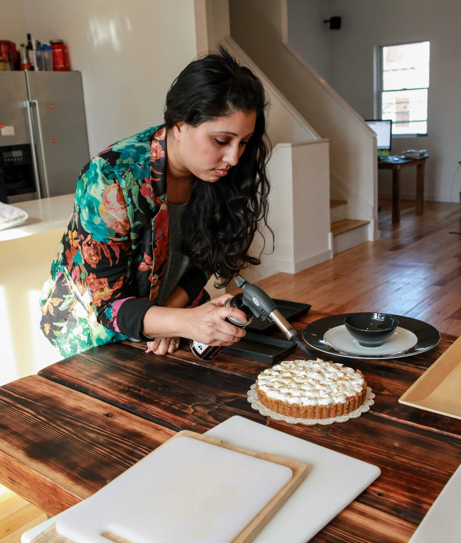 Chef Aarti in a floral jacket torching the top of a meringue tart in her kitchen.